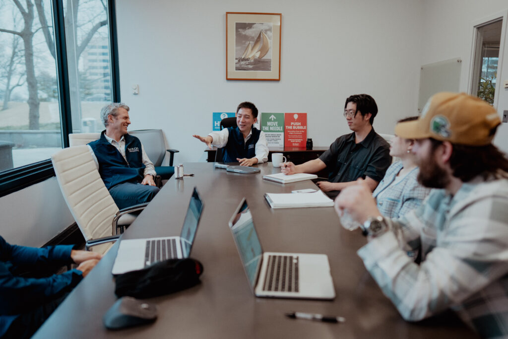 Linton team gathered around a conference table for a collaborative client meeting, with two members in branded navy vests leading the discussion alongside colleagues taking notes on laptops, set against a backdrop of branded posters reading "Move the Needle" and "Push the Bubble."