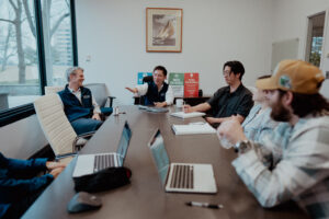 Linton team gathered around a conference table for a collaborative client meeting, with two members in branded navy vests leading the discussion alongside colleagues taking notes on laptops, set against a backdrop of branded posters reading "Move the Needle" and "Push the Bubble."
