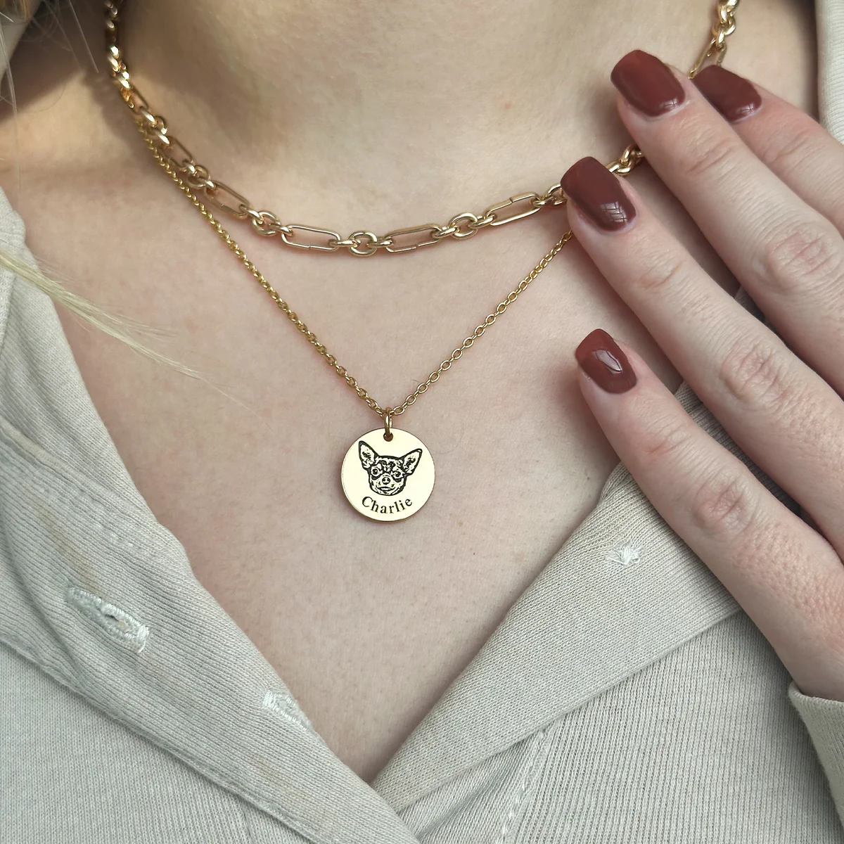 Close-up of a person wearing two layered gold necklaces: a chunky paperclip-style chain choker and a longer delicate chain with a round gold pendant engraved with a chihuahua's face and the name 'Charlie.' The person has dark red manicured nails and is wearing a beige textured top.