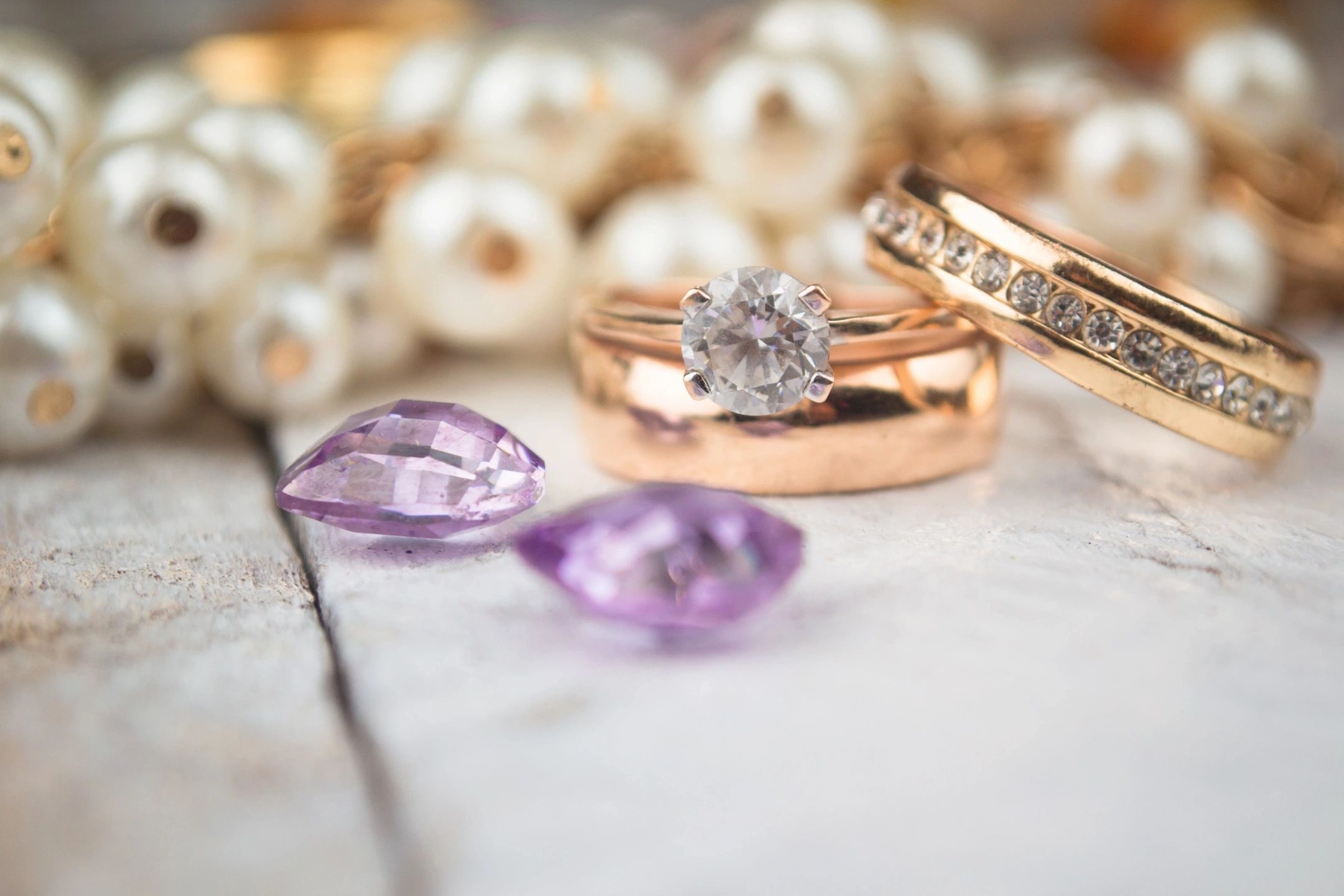 Close-up of fine jewelry on a rustic white wooden surface: a rose gold engagement ring with a round diamond, a gold wedding band with channel-set diamonds, two loose marquise-cut amethyst gemstones, and a strand of white pearls in the background