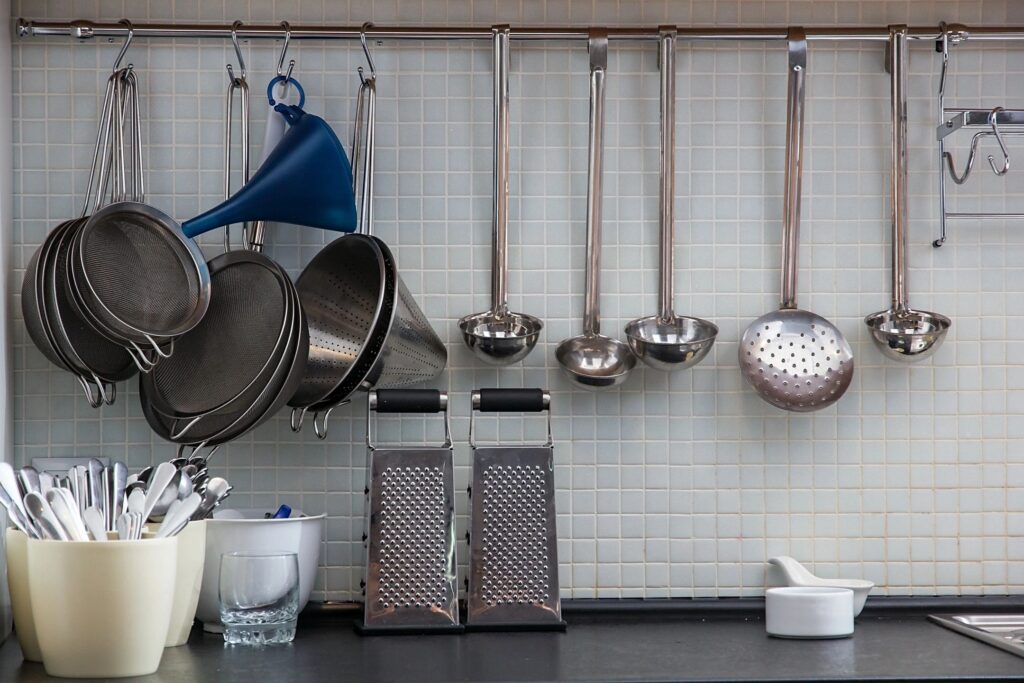 Kitchen utensils and cooking tools hanging on a wall rail, including metal strainers, ladles, graters, a funnel, and serving spoons above a countertop.