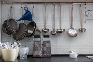 Kitchen utensils and cooking tools hanging on a wall rail, including metal strainers, ladles, graters, a funnel, and serving spoons above a countertop.