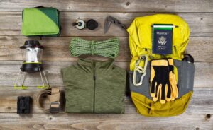 Hiking and travel gear arranged on a wooden surface, including a backpack, lantern, rope, jacket, gloves, passport, compass, binoculars, and outdoor tools.