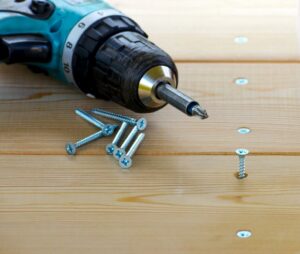 A blue power drill laying on a wooden table with loose screws scattered around
